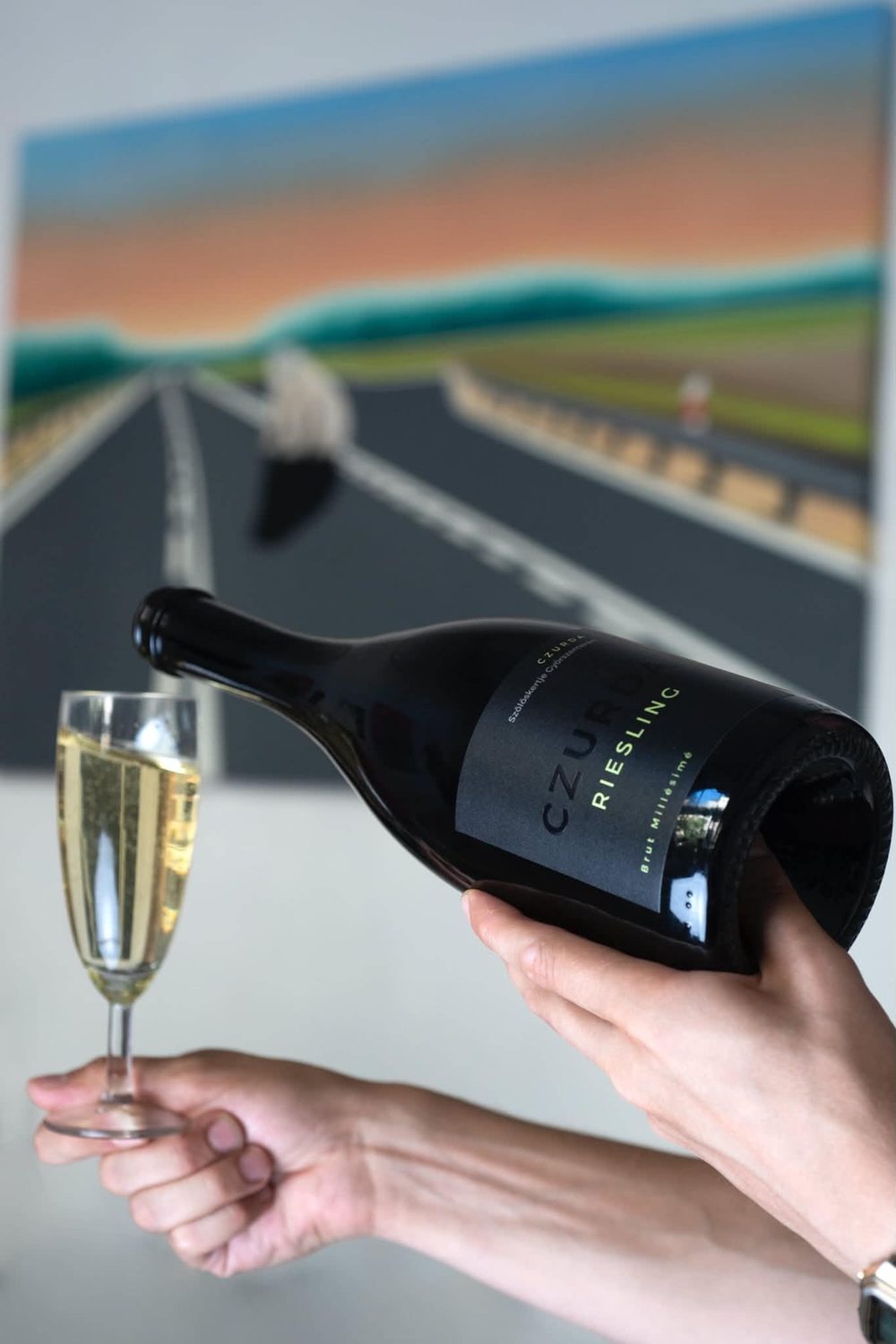 Egy Czurda Riesling Brut pezsgő palackja és egy pohár egy elmosódó, naplementés autópályát ábrázoló festmény előtt